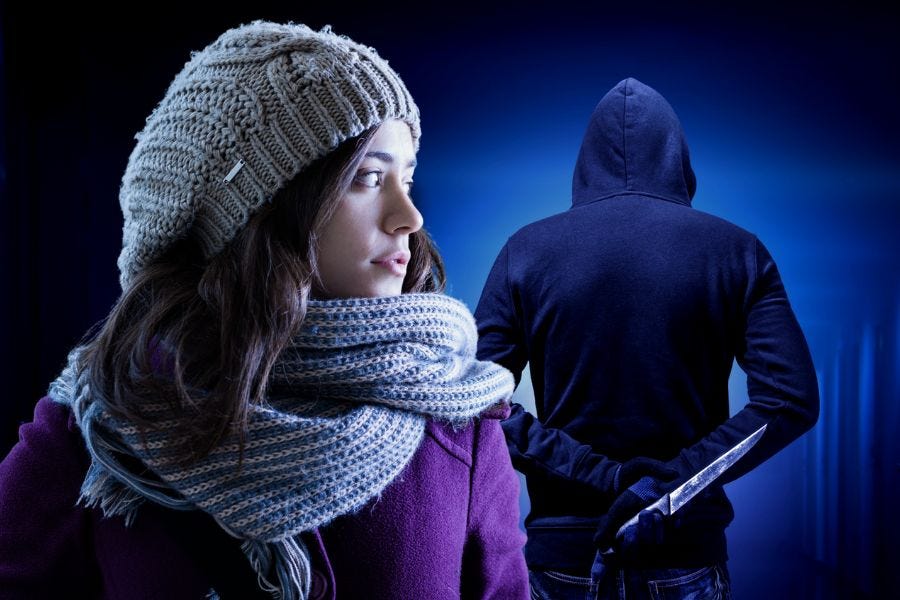 A girl in winter clothing looks nervously over her shoulder at a shadowy stalker in the background. He is facing away and holding a knife.